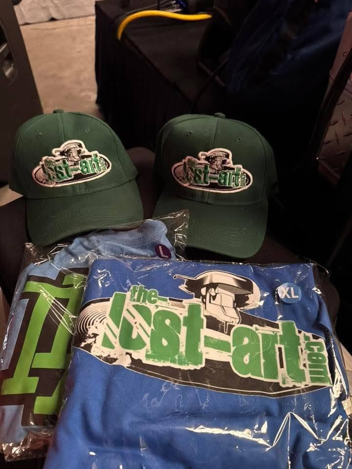 Two The-Lost-Art.com green embroidered caps and blue The-Lost-Art.com shirts laid out at the Florida underground show May 2026