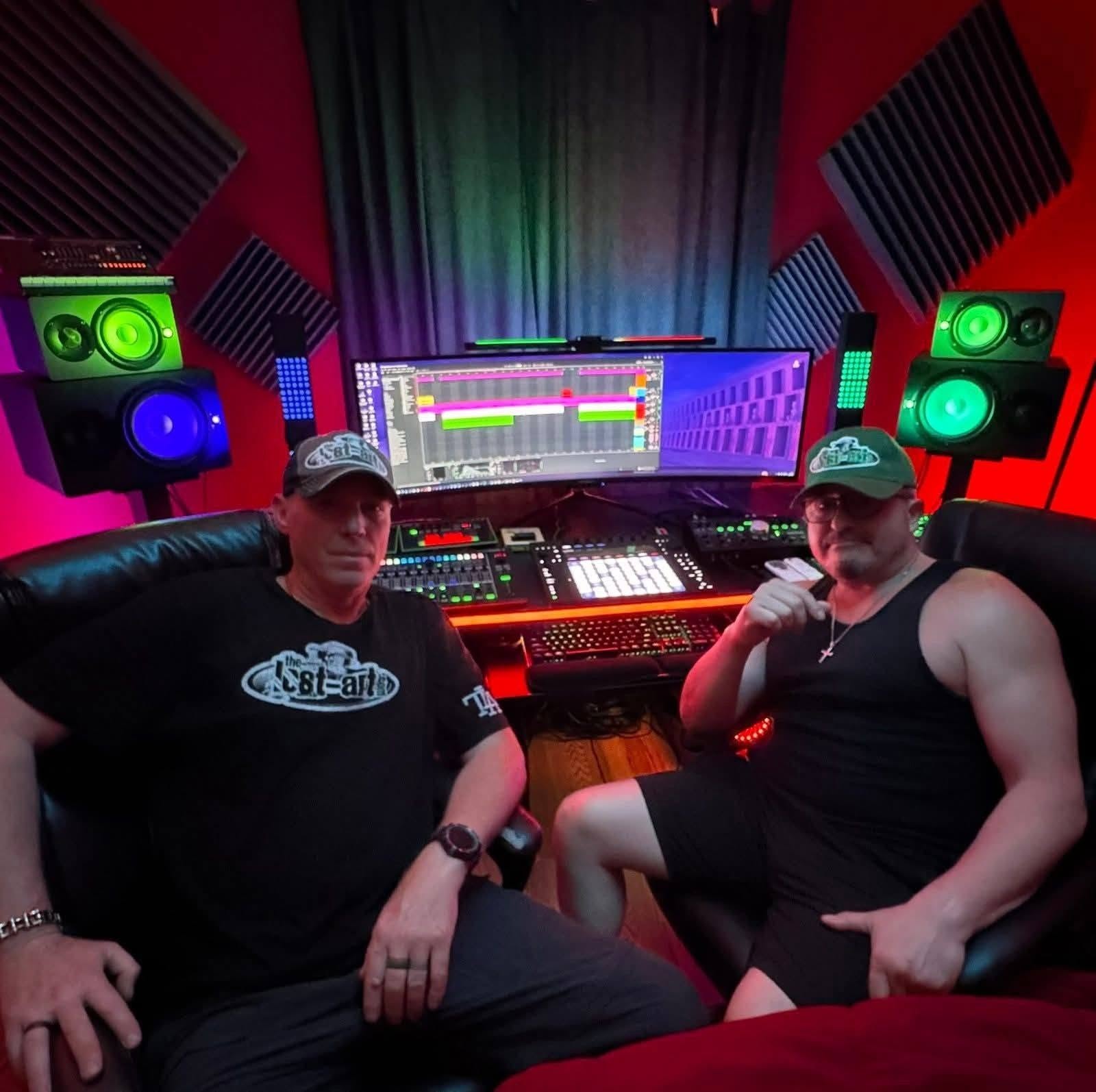 DJ Natural Nate® and a friend both wearing The-Lost-Art.com caps seated in front of a professional recording studio setup with green speakers and red lighting in Florida May 2026