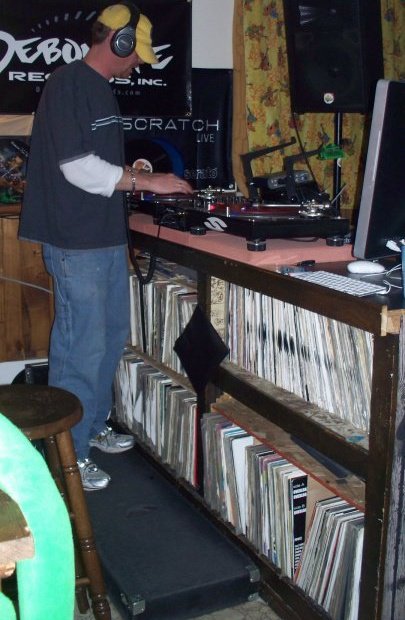 DJ Natural Nate® at the turntables, massive vinyl record collection on shelves below, Debonaire Records and Scratch Live banners behind him